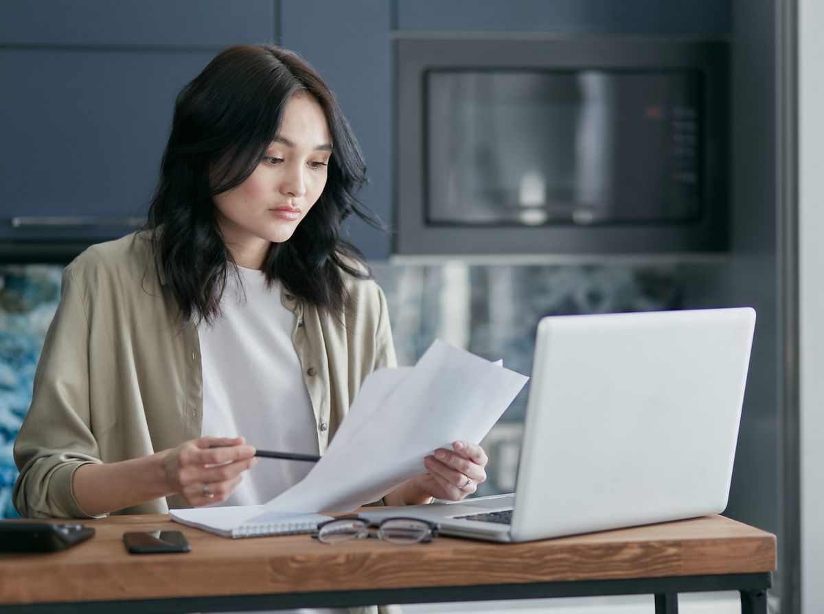 A woman reviewing paperwork beside her laptop at a home desk